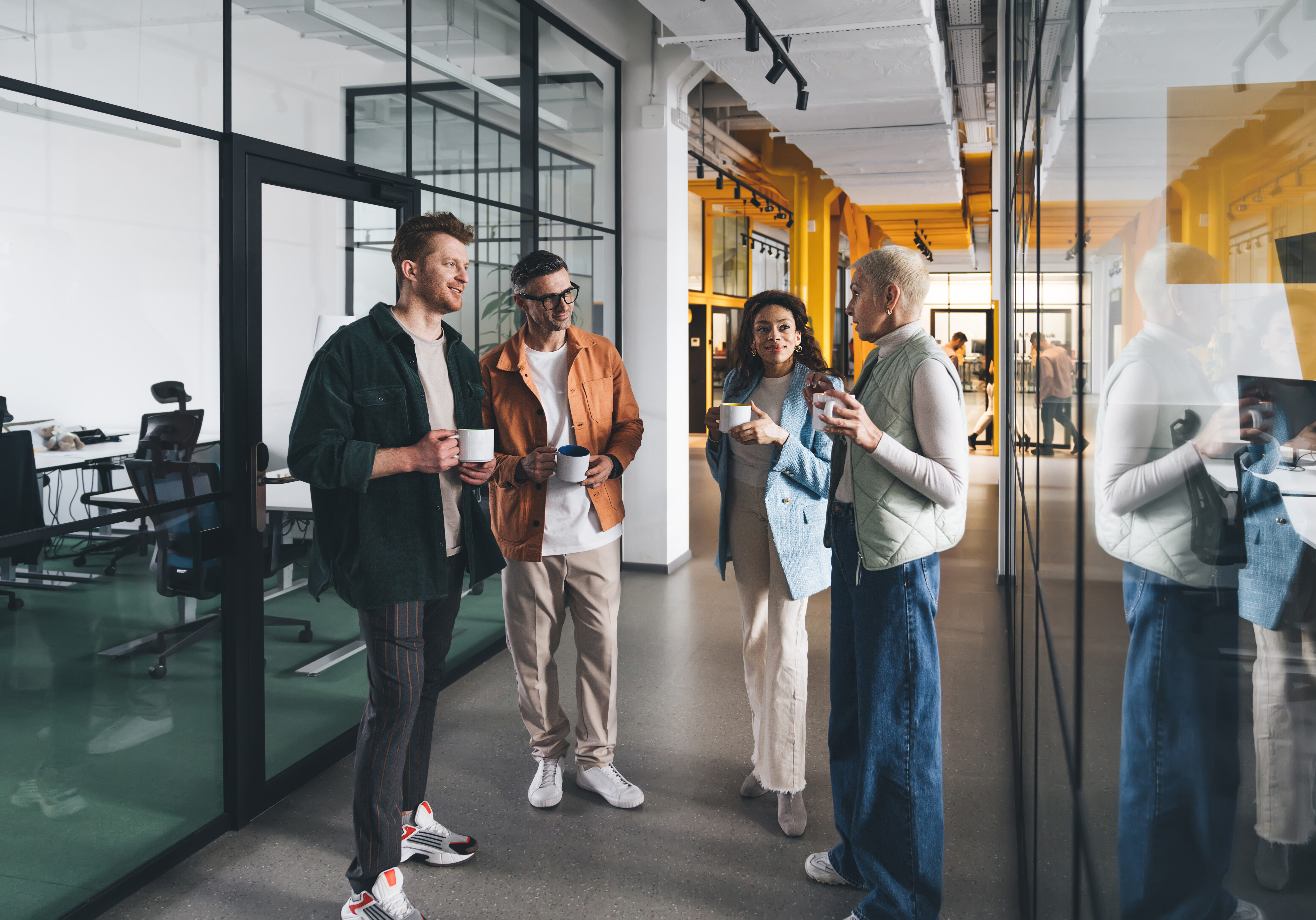 Group of multiracial team members wearing casual clothes standing in corridor of modern office and talking to each other with cups in hands during coffee break