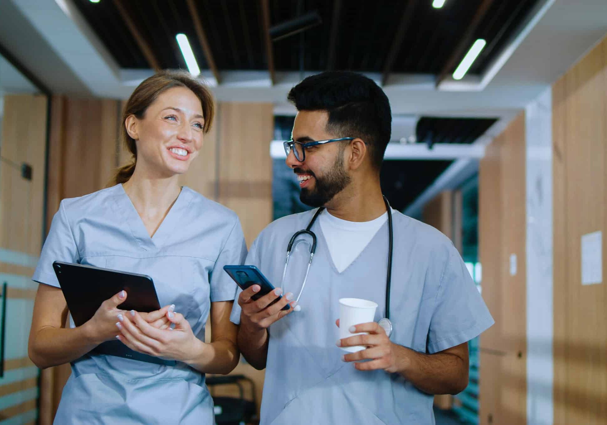 Portrait of skilled happy doctors walking in modern hospital hallways smiling and talking about their jobs. Caucasian doctor and young cheerful male Indian nurse having coffee.